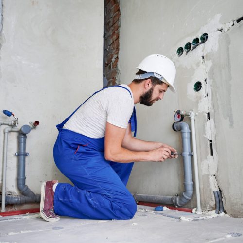Horizontal snapshot of young plumber working with grey sewer pipes, fixing them to wall with a help of screwdriver. Side view of plumber standing on knees wearing blue uniform and white helmet